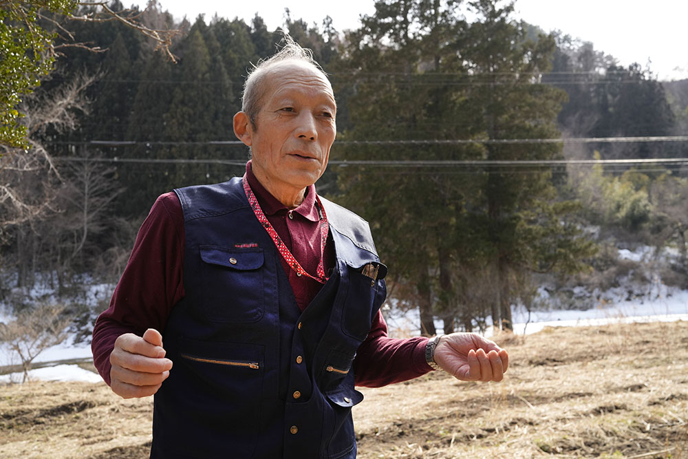 自宅と畜舎の跡地に立つ松本信夫。この場所に来ると、牛の殺処分が去年の出来事のように感じるという=2025年3月、福島県葛尾村